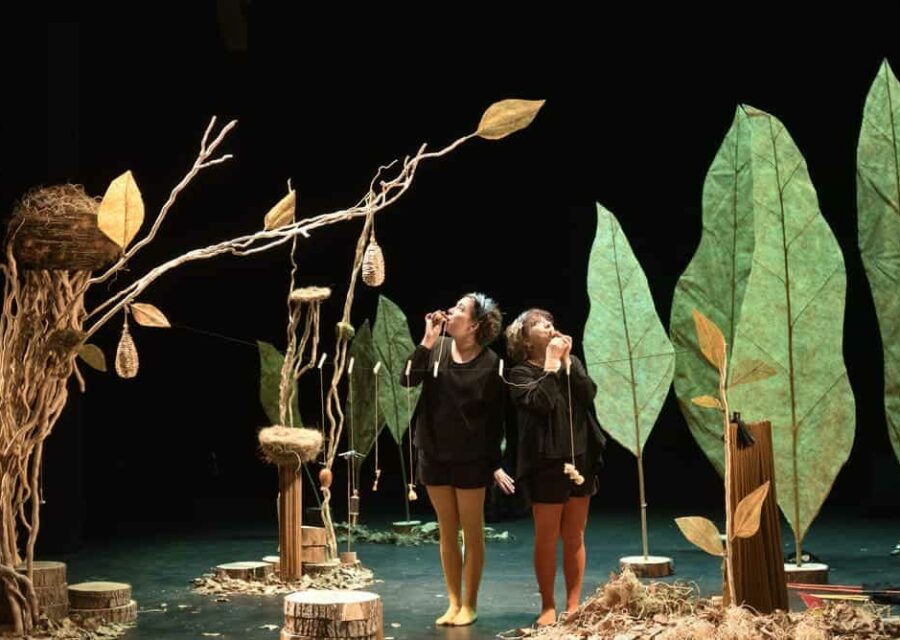 Dans un décor représentant des arbres avec des nids d'oiseaux, deux comédiennes soufflent dans des appeaux pour imiter le chant des oiseaux.