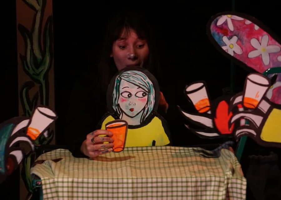 Une comédienne manipule une marionnette de papier représentant une petite fille à table avec un verre à la main.