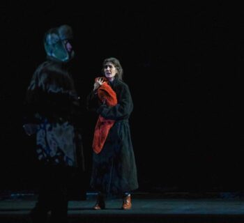 Une femme en manteau noir porte contre elle ce qui semble être un bébé emmailloté dans un châle rouge. Elle est tendue et regarde un homme en manteau noir et chapka.