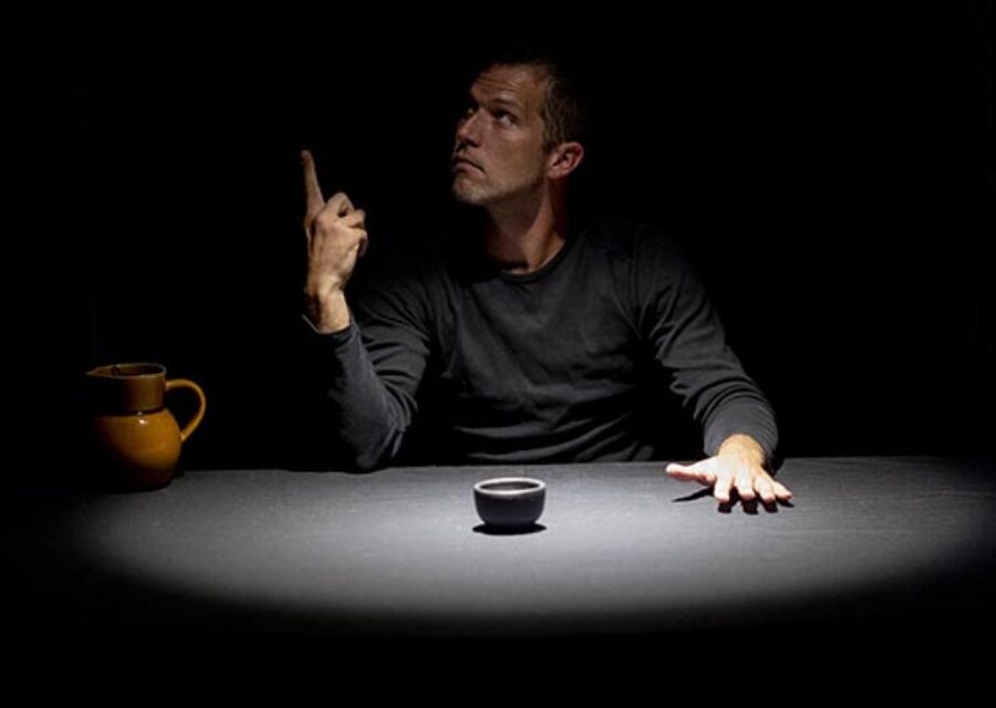 Le comédien-manipulateur est assis devant une table noire sur laquelle sont posés un petit bol au centre et une cruche à gauche. Sa main gauche est posée à plat sur la table devant lui. Il tend l'index de la main droite vers le haut et son regard suit cette direction.
