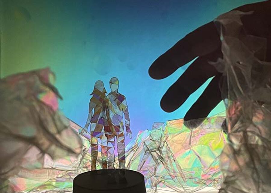 Deux silhouettes, un homme et une femme, posées sur un socle et entourées de matières colorés et transparentes. L'ombre d'une main géante apparaît à droite de l'image.