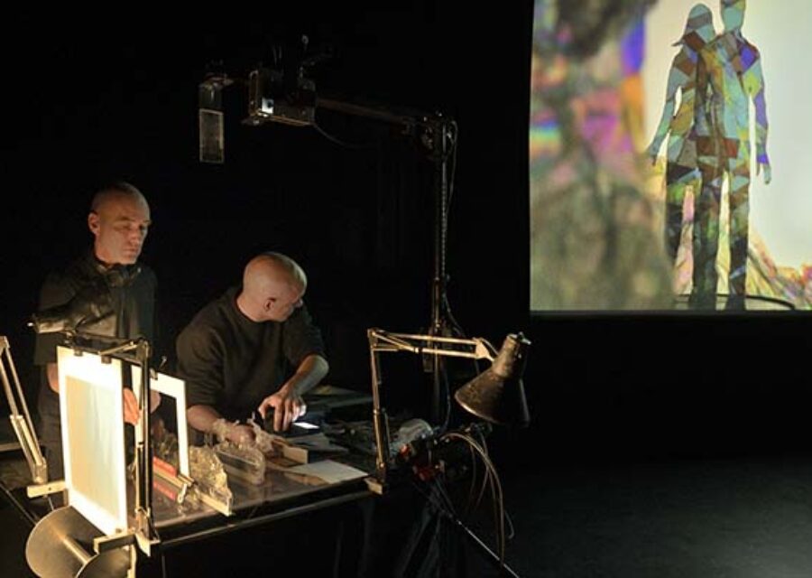 A gauche, sur une table, deux artistes manipulateurs disposent différents éléments de papier (personnages, matière, décors) dans un petit théâtre d'ombres dont l'image finale est projetée sur grand écran à droite de l'image. Ici, deux silhouettes (un homme et une femme) de plusieurs couleurs comme dans un kaléidoscope.