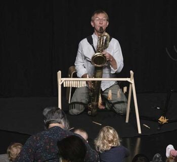 Des enfants et des adultes assis sur des coussins devant un musicien de face jouant du saxophone baryton. Il a devant lui un petit portique en bois sur lequel sont accrochés des anches et des bec de saxophone.