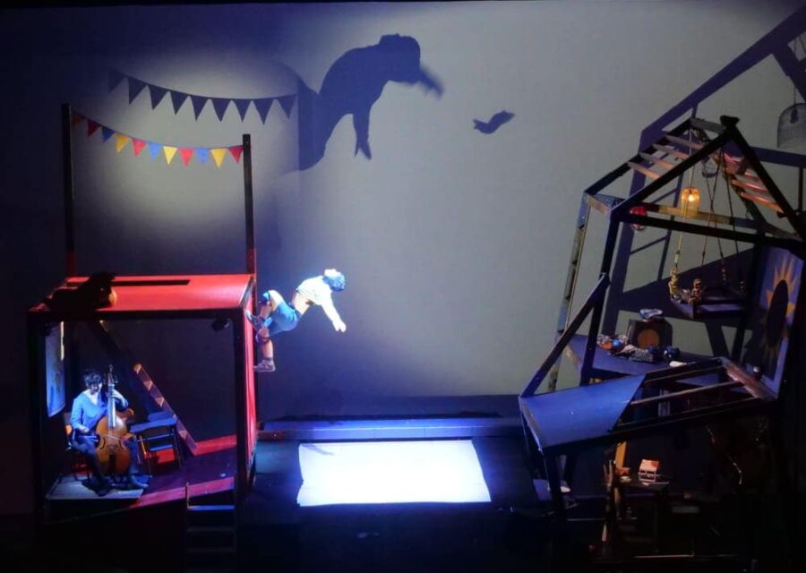 Vue générale du décor avec à gauche la structure représentant l'école et à droite , la maison bancale. Au milieu se trouve une toile de trampoline sur laquelle l'enfant vient de rebondir. Il est en suspension et son ombre se projette sur le fond de la scène.