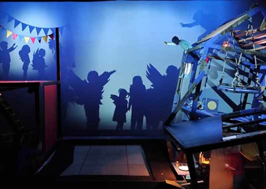 Vue générale du décor avec à gauche la structure représentant l'école et à droite , la maison bancale. L'enfant est dans la maison et fait signe à des enfants avec des ailes dont on ne voit que les ombres projetées sur la fond de la scène.