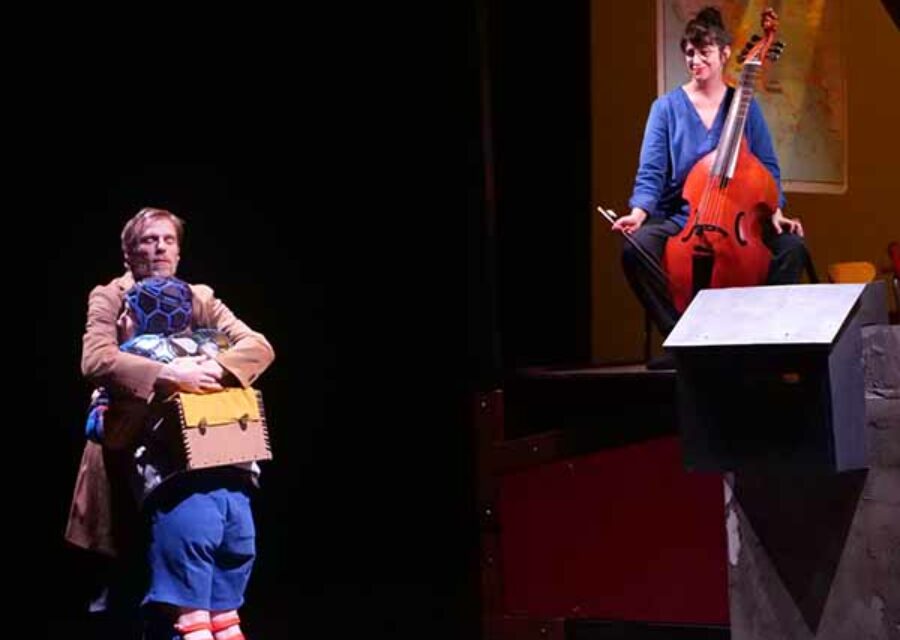 Un homme debout (le père) serre son fils dans ses bras. A droite et en surplomb, une violoncelliste joue.