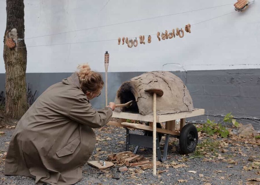 L'artiste fait cuire des petits pains dans un petits four en terre. Au-dessus d'elle, les petits-pains cuits sont suspendus sur un fil.