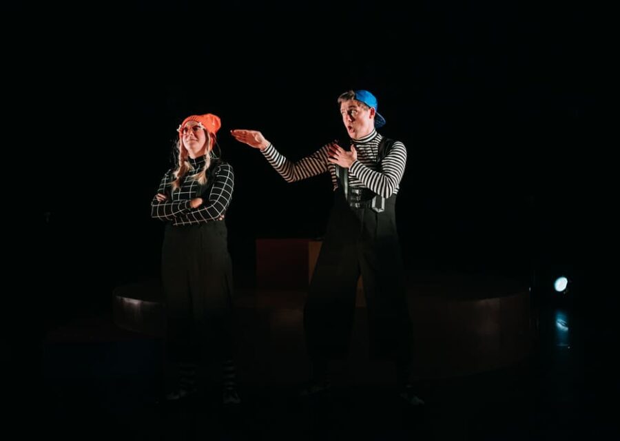 Une comédienne avec un pantalon noire, un haut noir quadrillé de lignes blanches, un bonnet rouge et des lunettes rouges excentriques. A côté d'elle, un comédien en pantalon noir, haut à rayures noires et blanches et casquette bleue portée à l'envers, tends le bras vers la comédienne qui lui tourne le dos..