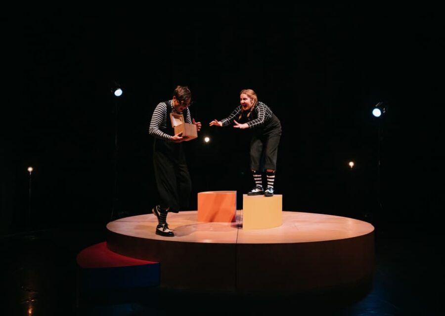 Sur une estrade circulaire, deux interprètes en costume noir avec des rayures blanches se disputent un coffret en bois.