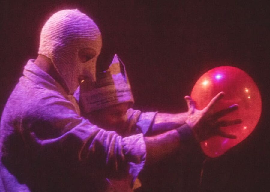 Un homme masqué tient un ballon de baudruche rouge entre ses mains tendues devant lui. Un enfant avec une couronne en papier journal est debout entre ses bras.