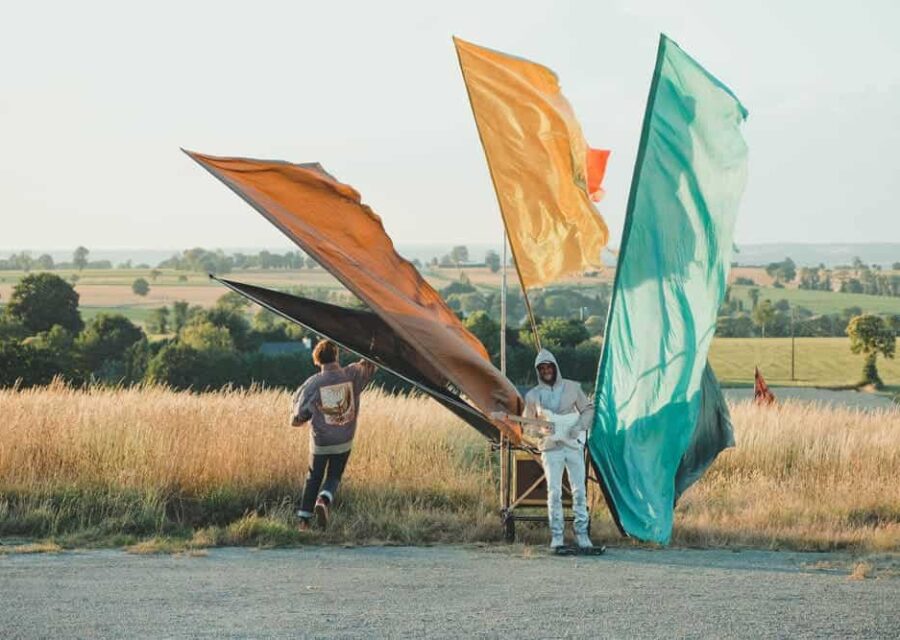 Photos du spectacle en extérieur avec le musicien jouant de la guitare électrique entouré de 4 étendards de couleur différente : noir, orangé, jaune, vert. Le comédien est visible de dos à gauche du musicien.