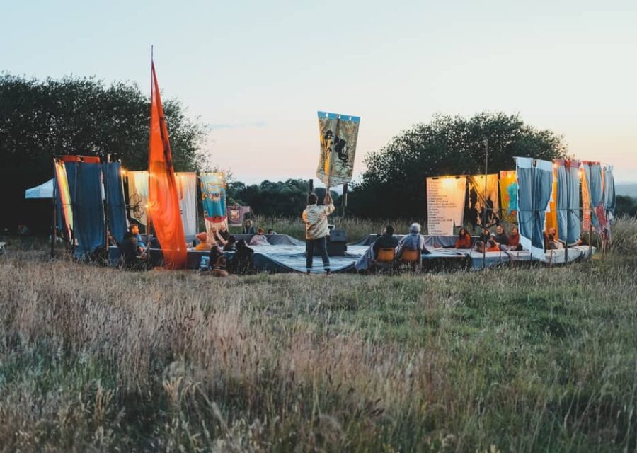 Photo du spectacle en extérieur à la tombée de la nuit. Le public est assis dans une arène circulaire entourée d'une quinzaine de bannières textiles qui font figure de tableaux. Le comédiens est au centre de dos et portant une bannière.