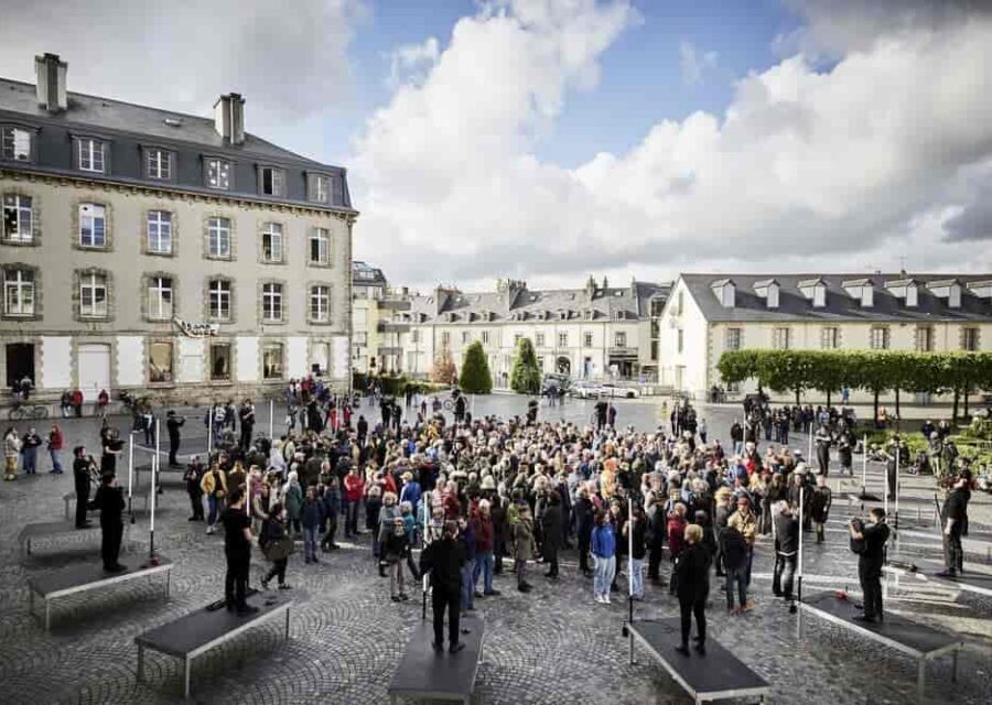 Les 20 sonneurs installés en rond sur une place avec le public debout au milieu.