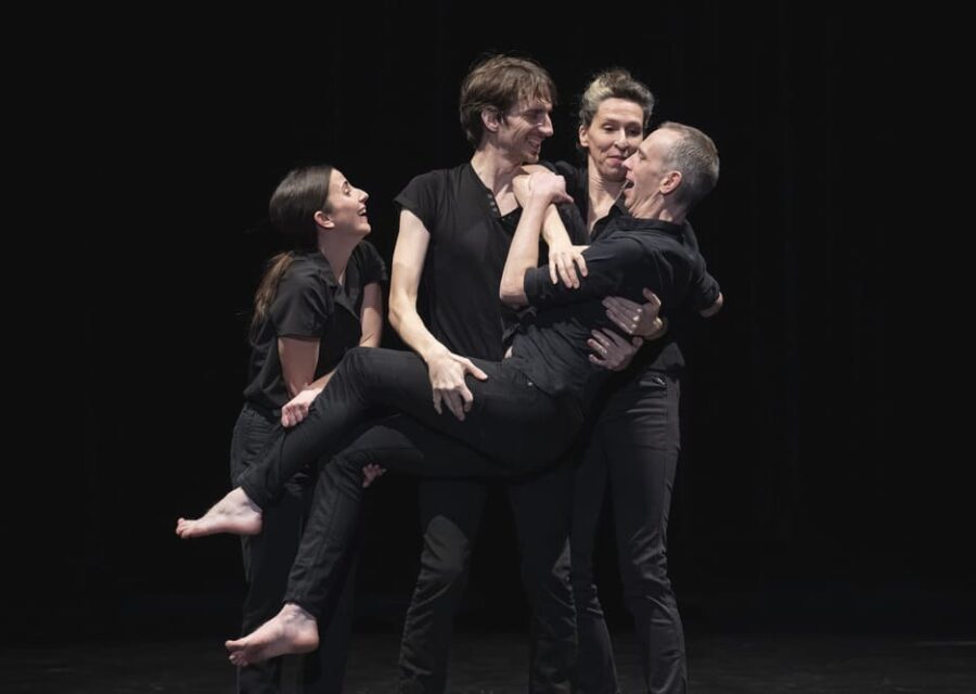 Un danseur entouré de deux danseuses portent un autre danseur presque allongé, sur le point de tomber. Ils sont tous habillés en jean et chemises noires et semblent s'amuser.