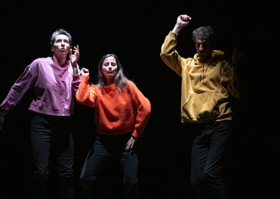 Deux danseuses et un danseur de face dansent comme en boîte de nuit. Ils portent un jean noir et chacun un sweat ou pull de couleur très vive : violet, rouge, jaune.