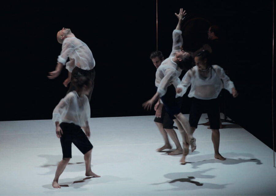 5 danseurs en bermuda noir et blouse blanche transparente ainsi qu'un musicien avec un gong en fond de scène. Deux des interprètes sont en l'air, en extension.