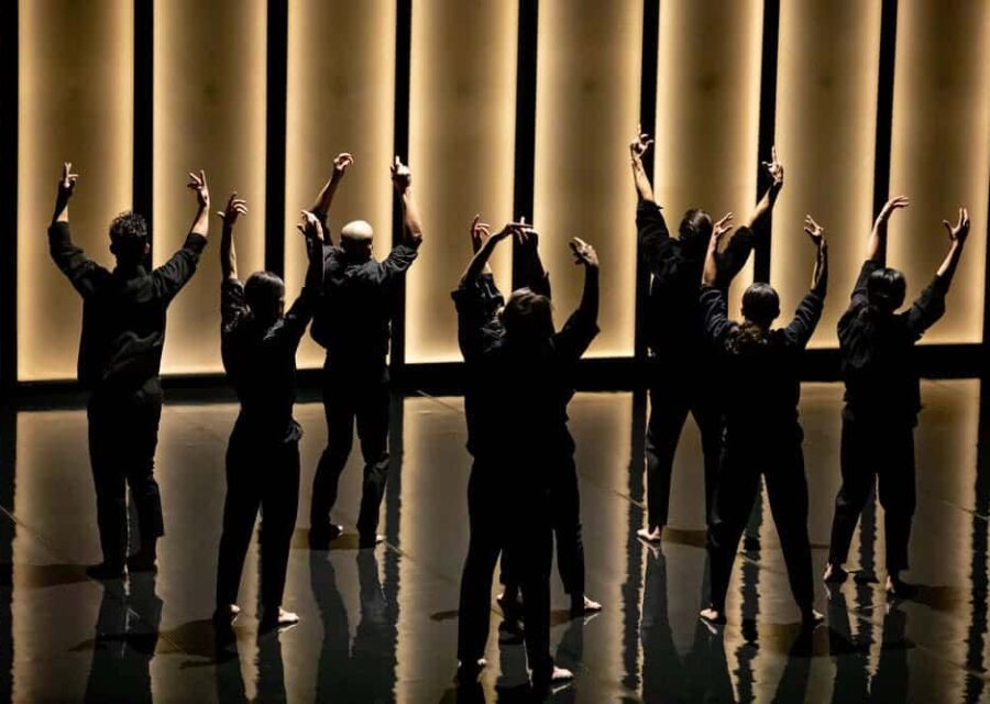 8 danseurs en ligne avec des pantalons et chemises noires, dans un décor contrasté entre noir et lumière.