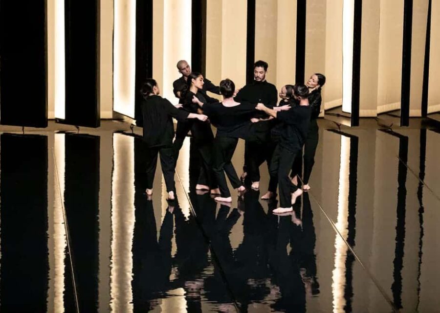 Groupe de 8 danseurs avec des pantalons et chemises noires, dans un décor contrasté entre noir et lumière.