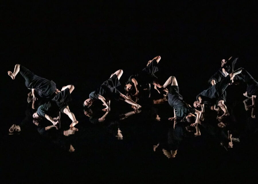 Les 8 danseurs sont en position tête au sol et une jambe en l'air. Leur image se reflète dans le sol à effet miroir.