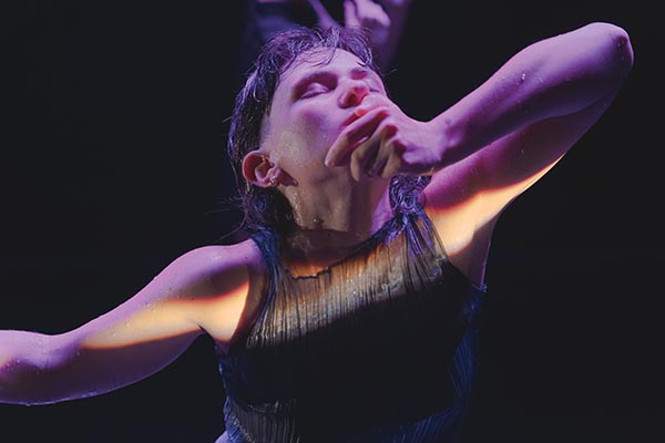 Gros plan sur le haut du corps d'une danseuse éclairé par un trait de lumière suivant la ligne de ses bras. Elle a le visage penché en arrière et les yeux fermés, sa main gauche est posée sur son menton.