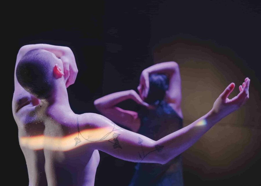 Au premier plan, le haut du corps nu d'une danseuse, de dos et un autre interprète en arrière plan.