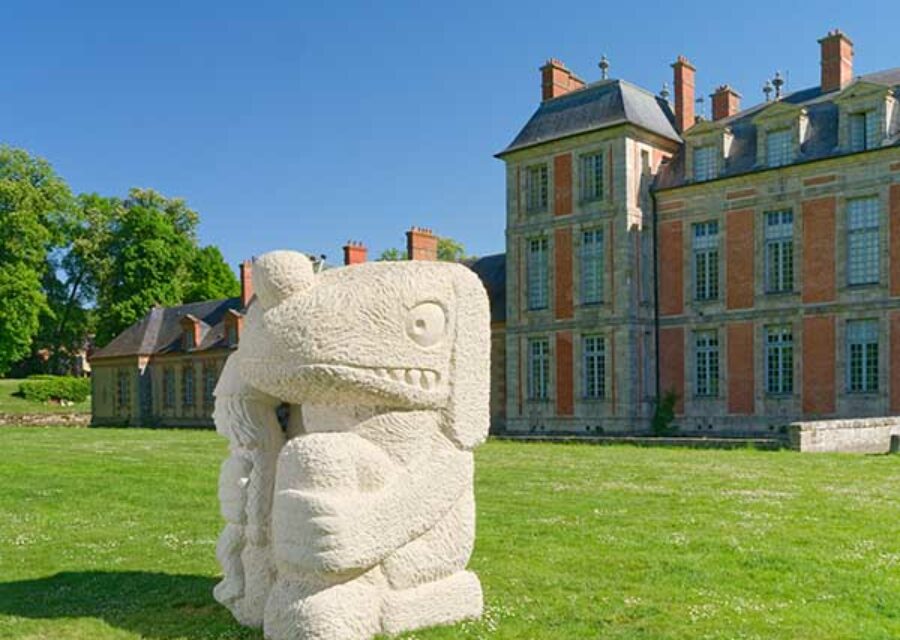 Sculpture intitulée Fate's fool, en grès de taille imposante sur la pelouse devant un château. L'oeuvre représente un personnage plutôt animal assis qui tient dans sa main droite un autre personnage humain plus petit.