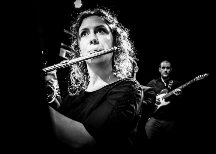 Photo en noir et blanc. Gros plan sur une musicienne qui joue de la flûte traversière et on voit en arrière plan un musicien qui joue de la guitare électrique.