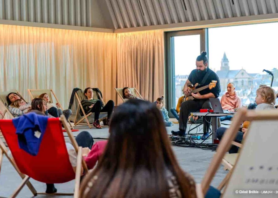 Le public est installé en cercle , assis dans des transats et on voit un musicien qui joue de la guitare électrique.