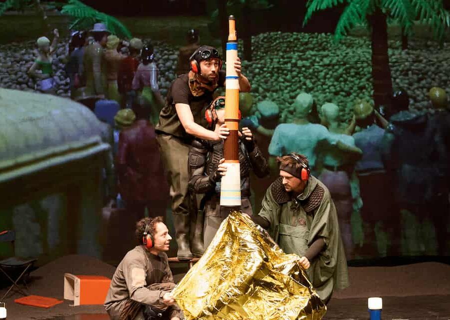 Reconstitution du lancement de la fusée Apollo 11 depuis Cap Canaveral avec assemblage de bouteilles thermos, lampe torche et couverture de survie sur fond de projection sur grand écran d'une foule constituée de petits personnages en plastique.