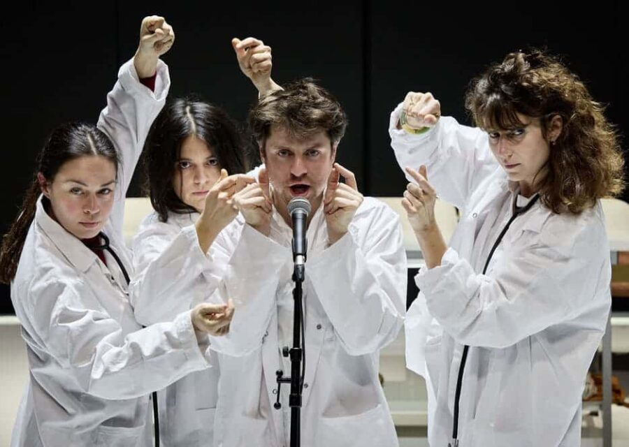 Un homme entouré de trois femmes chante dans un micro. Ils sont tous en blouse blanche et ont les bras en l'air. Ils claquent des doigts pour pulser le rythme.