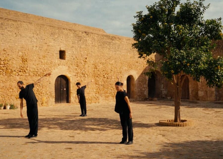 Trois danseurs habillés en noir et un arbre dans une cour entourée de hauts murs de couleur ocre.